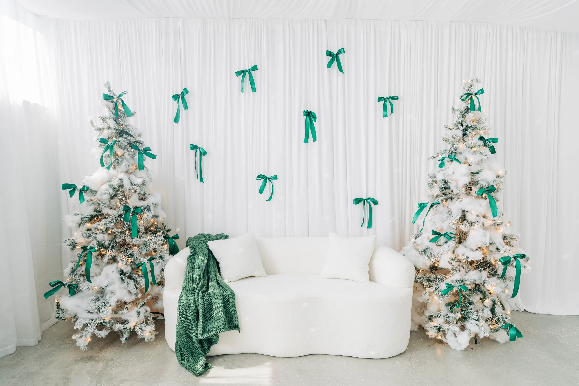 Two flocked Christmas trees and a curved white sofa against a draped backdrop scattered with green satin bows