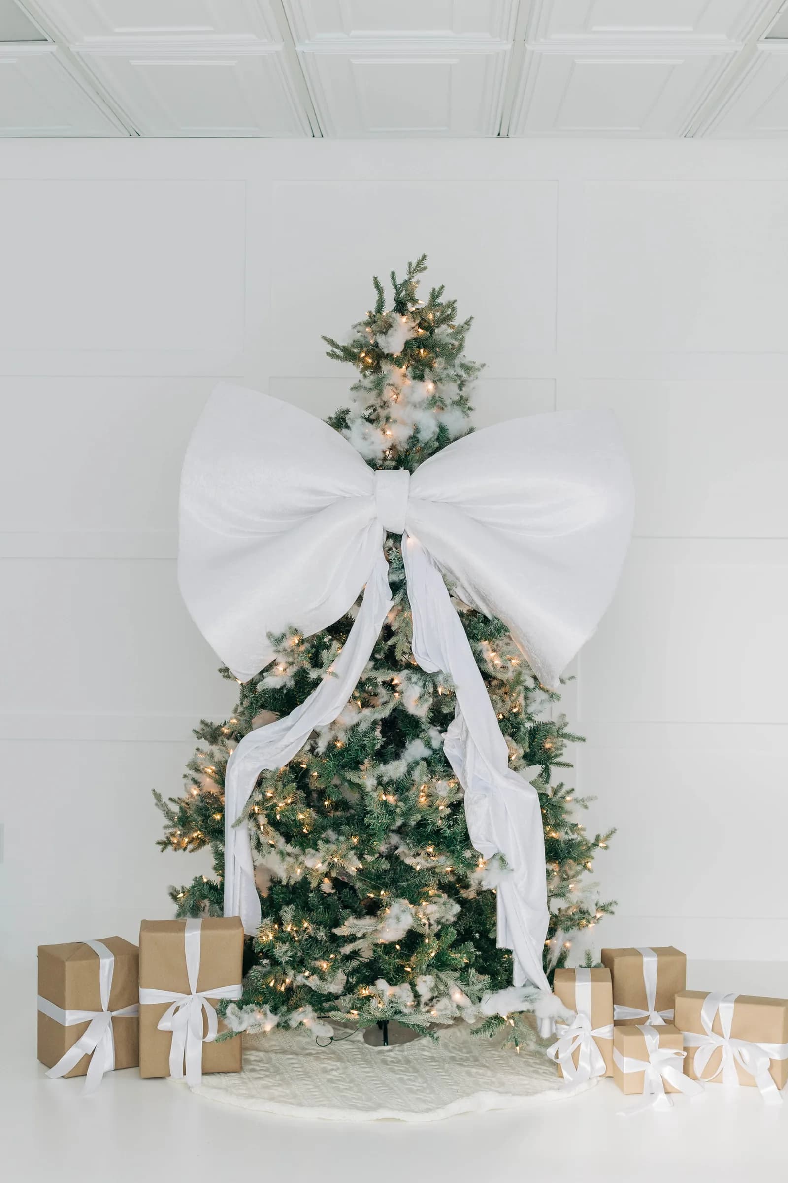A lit Christmas tree topped with an oversized white velvet bow, kraft paper presents tied in white ribbon at its base