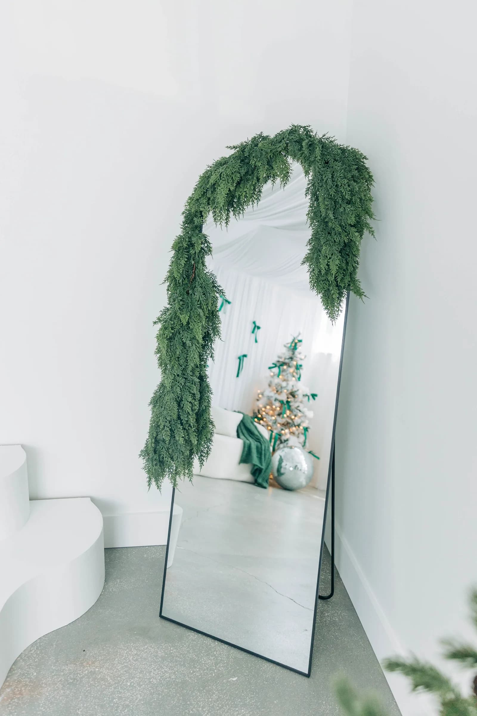 A full length mirror draped in cedar garland, reflecting the flocked trees and green bow backdrop across the room