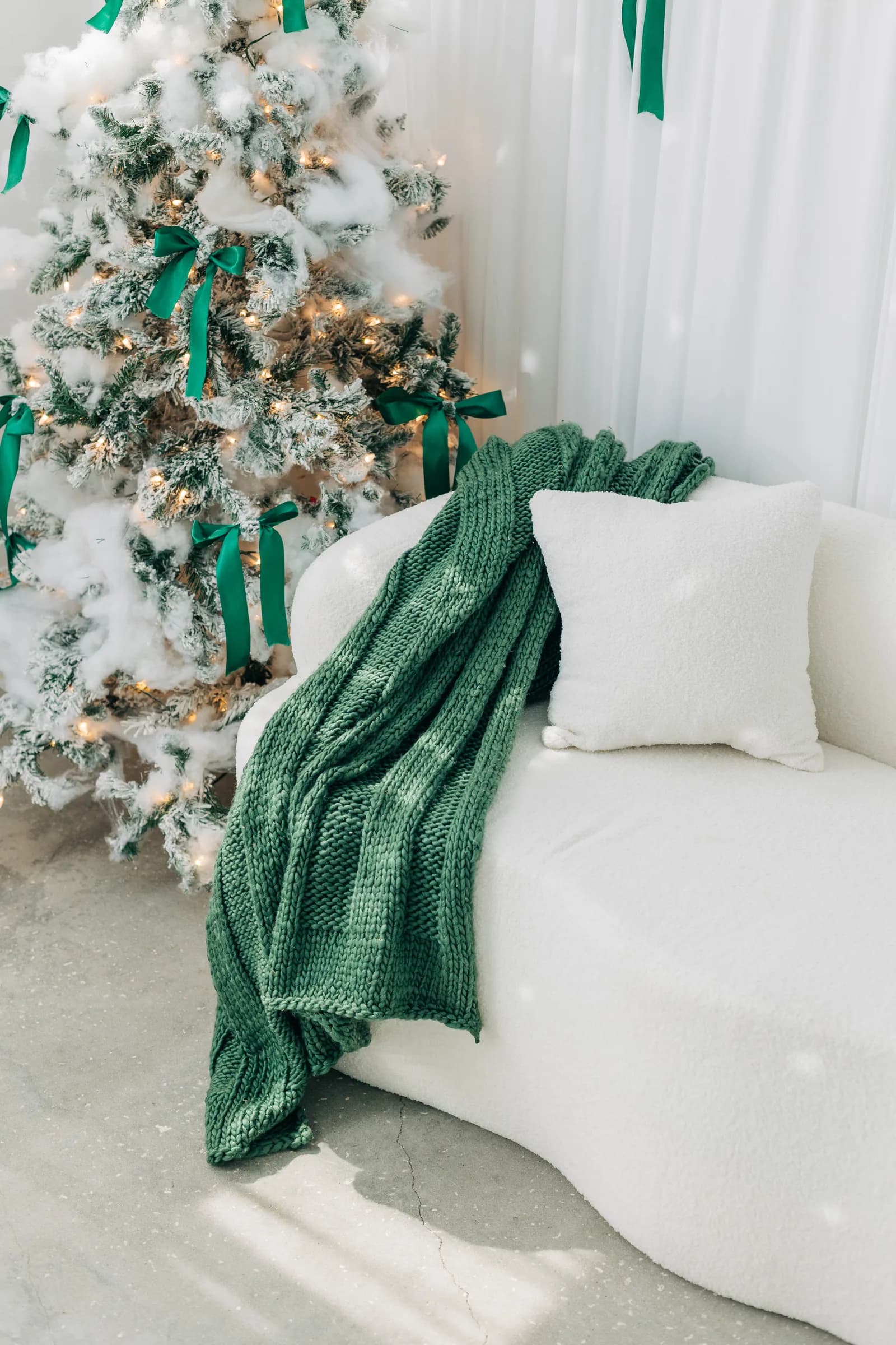 A sage knit throw over the curved white sofa beside a flocked tree tied with green ribbon