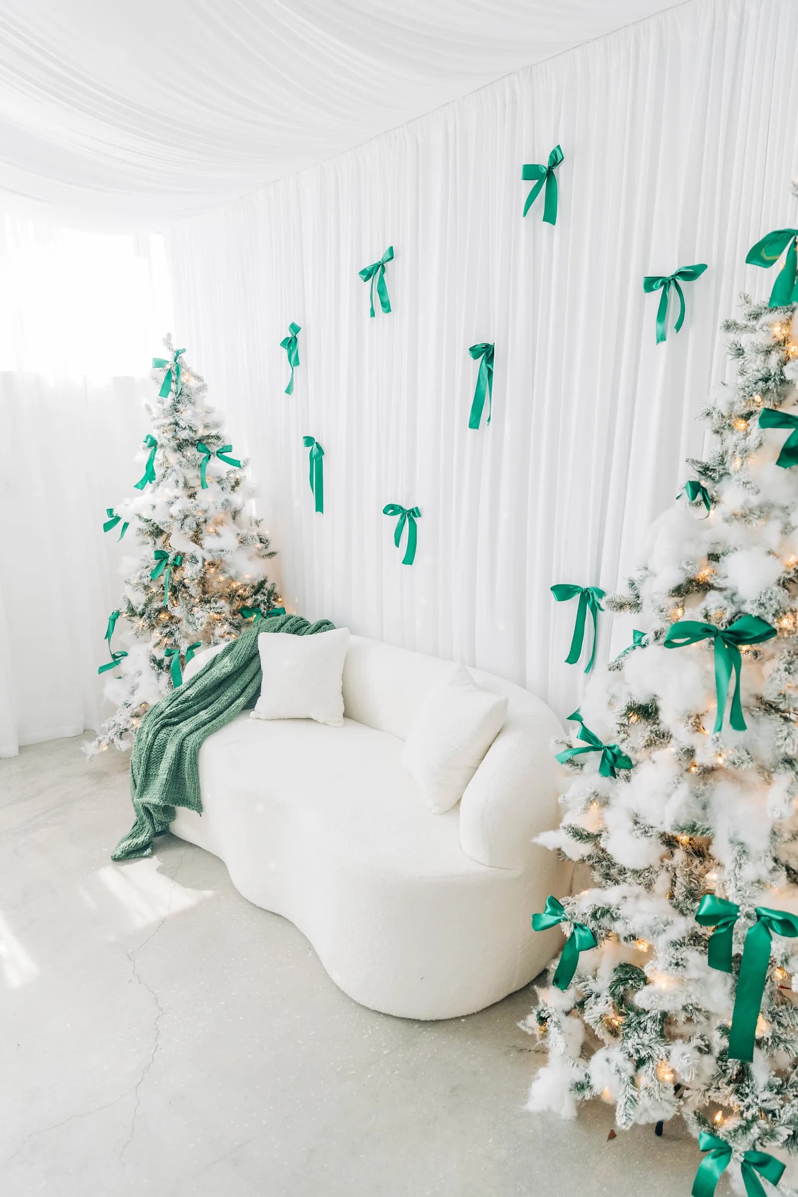 Two flocked Christmas trees and a curved white sofa in front of a draped backdrop scattered with green satin bows