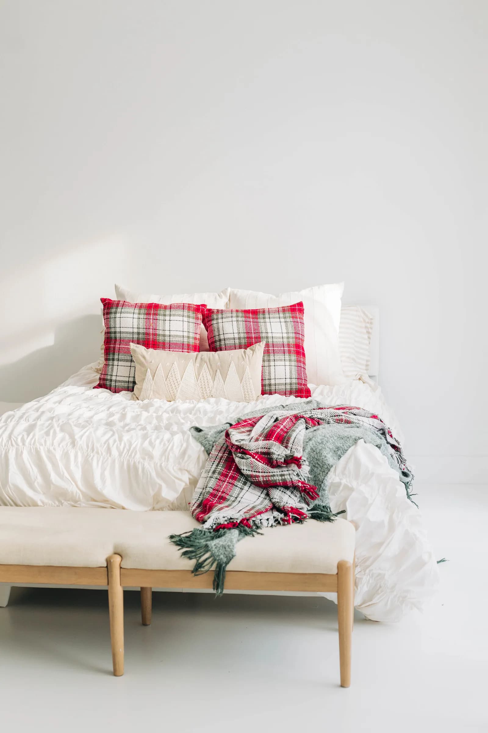 A studio bed made up in white with red plaid pillows and a fringed plaid throw across the wooden bench at its foot