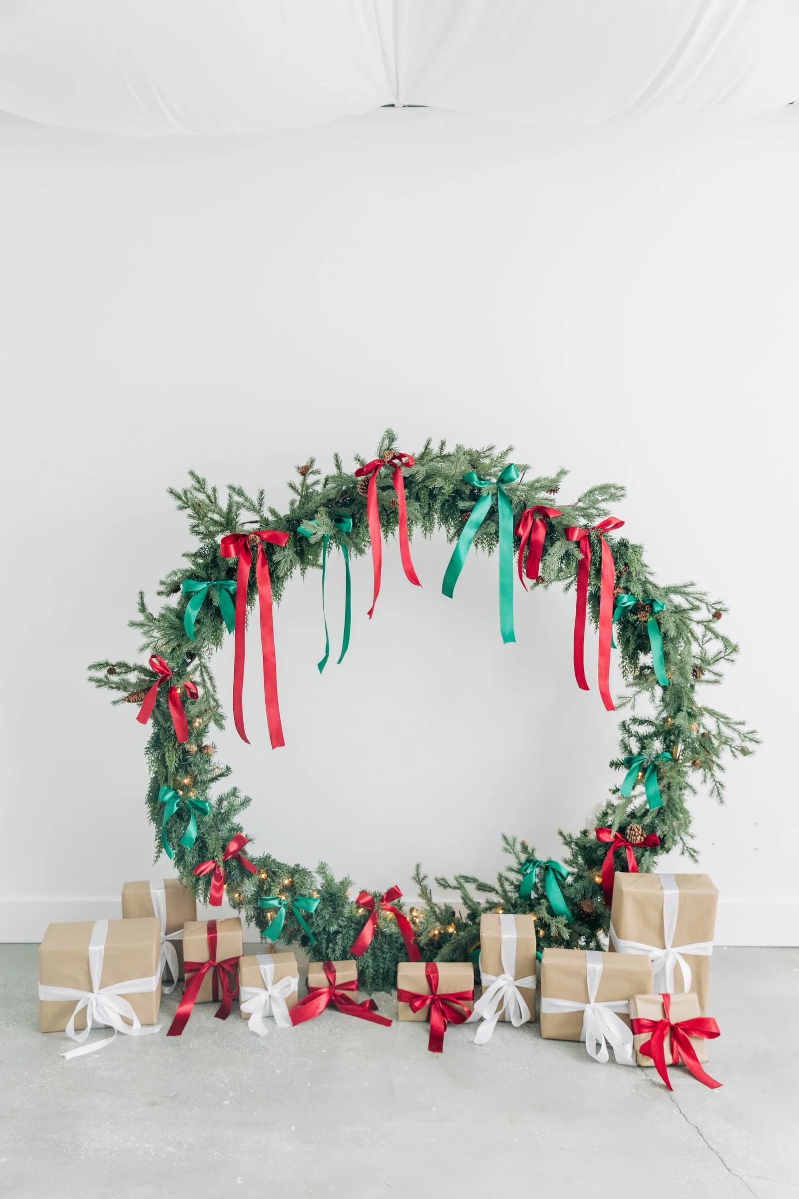 An oversized circular greenery wreath tied with red and green satin ribbon, wrapped presents on the concrete floor beneath it